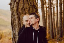 Sonriente pareja homosexual abrazando y mirando a la cámara cerca del árbol en el bosque y pintoresca vista del valle - foto de stock
