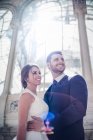 Знизу молодий елегантний чоловік приймає жінку у весільній сукні біля ретро-палацу з багатьма вікнами в сонячний день — стокове фото