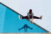 Бородатый парень в модном наряде, прыгающий вверх и смотрящий в камеру на голубую стену современного здания в солнечный день — стоковое фото