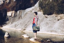 Женщина, стоящая на скале с футляром рядом с чистой водой в озере — стоковое фото