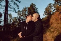 Casal homossexual abraçando e beijando na passarela na floresta em dia ensolarado no fundo borrado — Fotografia de Stock