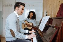 Vista lateral del joven tocando el piano cerca de la mujer negra tocando la guitarra en el estudio de música - foto de stock