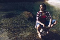 Felice donna adulta seduta sulla roccia in tranquilla acqua trasparente del lago godendo la natura e sorridendo via — Foto stock