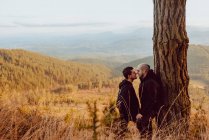 Cheerful homosexual couple standing and looking at each other near tree in mountains — Stock Photo