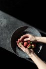 Mãos cortadas de pessoa segurando pilha de morangos frescos perto de tigela no fundo escuro — Fotografia de Stock