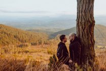 Heureux couple homosexuel debout et regardant l'autre près de l'arbre dans les montagnes — Photo de stock