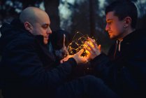 Homosexual couple with illuminated fairy lights sitting in murk forest in evening — Stock Photo