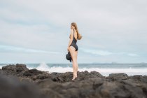 Вид сбоку привлекательной женщины в купальнике, держащей шляпу и смотрящей в камеру, стоя на каменистом берегу у волнистого моря на фоне пасмурного неба — стоковое фото