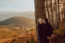 Side view of cheerful homosexual couple embracing and looking at each other near tree in forest and picturesque view of valley — Stock Photo