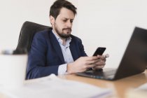 Konzentrierter junger Mann spricht am Handy und surft am Laptop am Tisch im Büro — Stockfoto