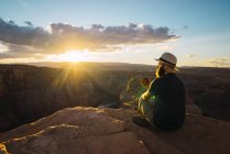 Side view of bearded guy in hat enjoying hot beverage and looking at setting sun while sitting near wonderful canyon on West Coast of USA — Stock Photo