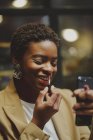 Africano americano elegante mulher colocando batom nos lábios e segurando telefone celular na rua — Fotografia de Stock