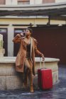 Africano americano mulher elegante com bagagem falando no telefone celular e de pé perto de monumento na rua — Fotografia de Stock