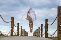 Back view of ballerina in dress with white veil in air on footbridge under blue sky in sunny day — Stock Photo