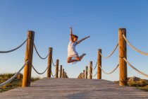 Молодая балерина в белом с поднятой ногой в воздухе на пешеходном мосту и голубым небом в солнечный день — стоковое фото