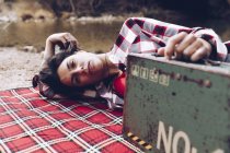 Sorrindo mulher adulta deitada em xadrez na margem do lago em falésias segurando velho caso enferrujado com provisão sorrindo para a câmera — Fotografia de Stock
