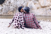 Погляд на молоду пару, що сидить і цілується на скелястому узбережжі гірської річки біля скелі. — стокове фото
