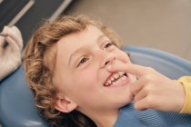 Веселий милий хлопчик вказує на зуб, лежачи на стоматологічному кріслі в сучасній клініці — стокове фото