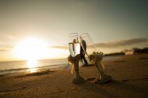 Крупные бокалы шампанского на песчаном побережье и молодожёны, обнимающиеся у воды на закате на размытом фоне — стоковое фото