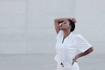 Sensual mujer étnica de pelo corto de moda en camisa blanca posando contra la pared gris - foto de stock