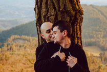 Alegre pareja homosexual abrazando cerca del árbol en el bosque y pintoresca vista del valle - foto de stock