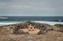 Stilvolle Frau in Badebekleidung und Hut sitzt in einem Steinkreis und meditiert, während sie an bewölkten Tagen Zeit in der Nähe des winkenden Meeres verbringt — Stockfoto