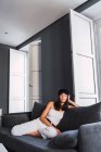 Приваблива молода стильна жінка в кепці дивиться на камеру, відпочиваючи на дивані біля балкона вдома — стокове фото