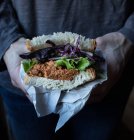 Couper les mains de la personne tenant le sandwich du pâté des tomates, la salade fraîche et le chou sur le plateau près du couteau sur la planche en bois — Photo de stock