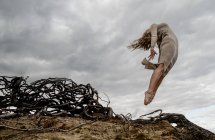 From below young ballerina in dress in air near dry branches and cloudy sky — Stock Photo
