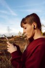 Jeune femme hipster avec perçage et écouteurs écoutant de la musique avec téléphone portable et marchant sur la route de campagne — Photo de stock