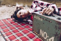 Smiling adult woman lying on plaid on lake shore in cliffs holding old rusty case with provision smiling at camera — Stock Photo