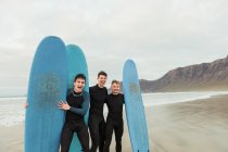 Drei junge Männer in schwarzen Badeanzügen lachen und blicken in die Kamera, während sie blaue Surfbretter in der Hand halten und am Ufer in der Nähe des Meeres stehen — Stockfoto