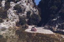 Vue arrière du jeune couple assis et embrassant sur la côte rocheuse de la rivière de montagne près de la falaise — Photo de stock