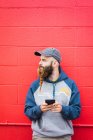Tipo atractivo con barba trenzada navegando por el teléfono inteligente mientras se inclina pared roja en la calle de la ciudad - foto de stock