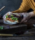 Mãos de colheita de pessoa que mantém o sanduíche de patê de tomates, salada fresca e repolho na bandeja perto de faca na tábua de madeira — Fotografia de Stock
