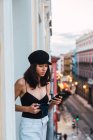 Joven mujer delgada en gorra con taza usando teléfono móvil y de pie en el balcón en la calle con luces en la noche - foto de stock