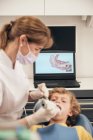 Mulher em máscara e uniforme médico fazendo varredura de dentes de menino enquanto trabalhava na clínica odontológica — Fotografia de Stock