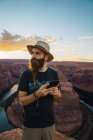 Человек в шляпе на мобильном телефоне, стоя на фоне каньона и реки во время заката на Западном побережье США — стоковое фото
