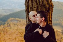 Alegre pareja homosexual abrazando y mirando a la cámara cerca del árbol en el bosque y pintoresca vista del valle - foto de stock