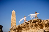 Снизу молодые загадочные женщины с поднятыми руками, держащие белый текстиль и позирующие на скалах и голубом небе — стоковое фото