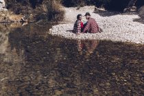 Vista laterale della giovane coppia seduta sulla costa rocciosa del fiume di montagna vicino alla scogliera — Foto stock