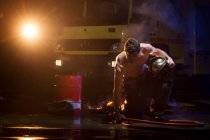 Shirtless muscular male in fireman uniform sitting on burning floor during dangerous mission — Stock Photo