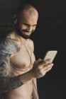 Bald shirtless athlete smiling and browsing smartphone while standing in dark gym and listening to music during workout — Stock Photo