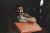 Красивий бородатий спортсмен з голим торсом посміхається і дивиться геть, сидячи на розмитому тлі тренажерного залу під час тренування — стокове фото