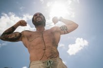 Мускулистый мужчина без рубашки сгибает бицепсы и показывает язык, стоя на фоне облачного неба во время тренировки на открытом воздухе — стоковое фото