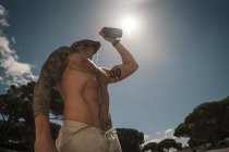 Мужчина без рубашки пьет пресную воду, стоя на фоне синего неба с ярким солнцем во время тренировки на открытом воздухе — стоковое фото