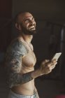 Красивый татуированный мужчина со смартфоном смеется и слушает музыку, стоя в современном спортзале во время тренировки — стоковое фото