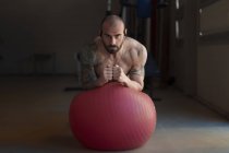 Schöner bärtiger Typ, der in die Kamera schaut, während er im Fitnessstudio Plank-Übungen auf Schweizer Ball durchführt — Stockfoto