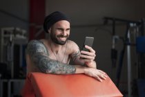 Atleta tatuado sin camisa riéndose con smartphone en el gimnasio - foto de stock