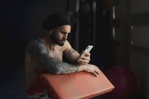 Atleta tatuado sin camisa con smartphone en el gimnasio - foto de stock